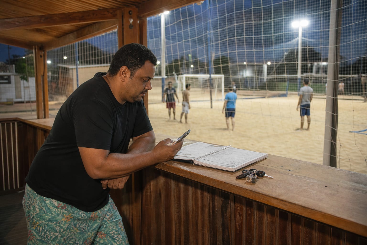 Dono de arena esportiva no balcão da recepção com caderno de agenda aberto e celular na mão, com a quadra iluminada ao fundo