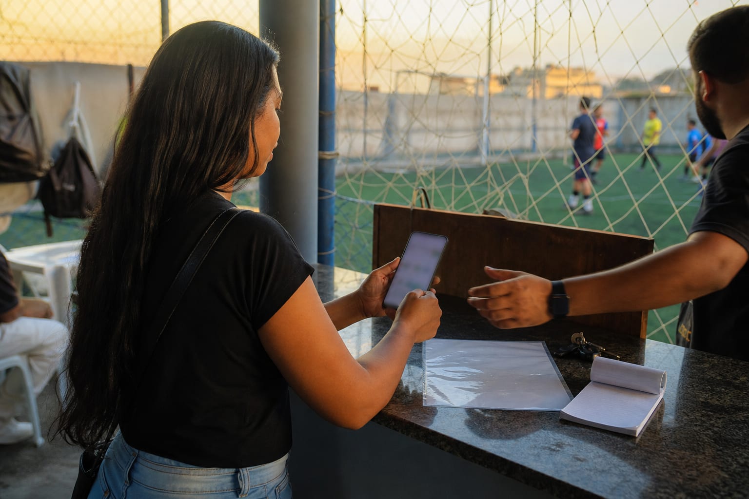 Mãos segurando celular com tela de pagamento no balcão de uma arena esportiva, com talão em branco sobre a bancada