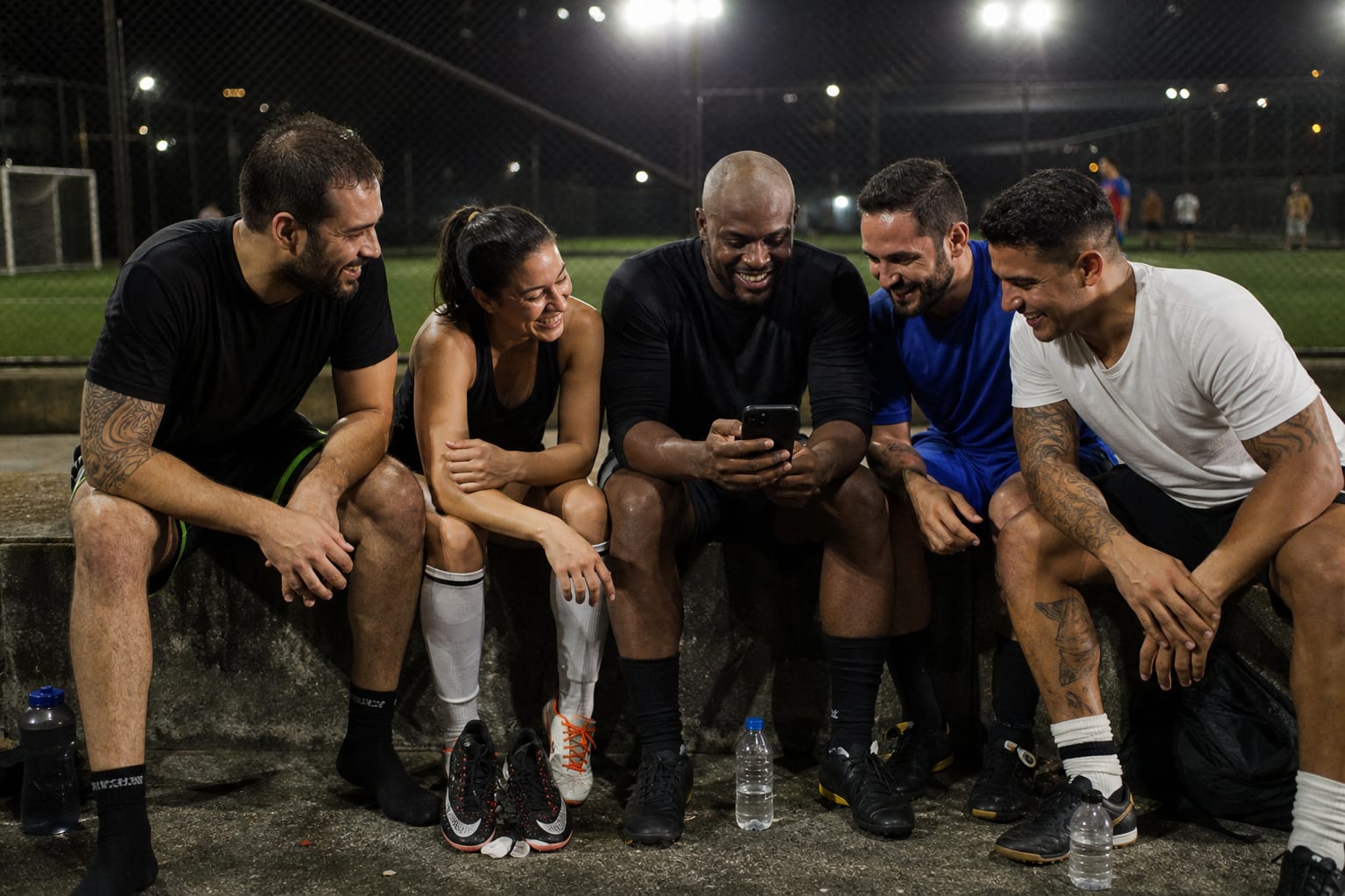 Grupo de amigos sentados ao lado da quadra de society depois do jogo, um deles mostrando o celular aos outros