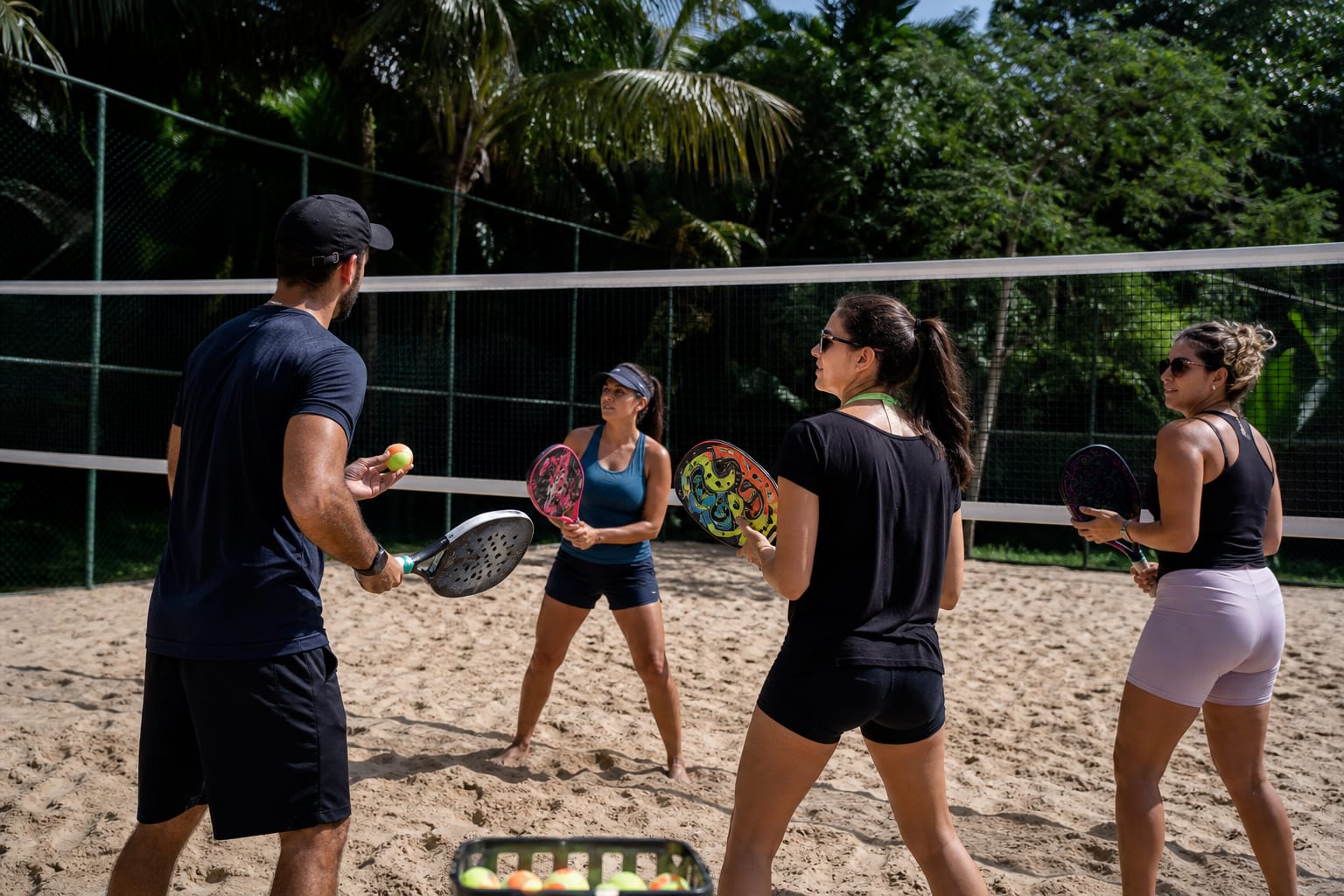 Professor dando aula de beach tennis para três alunas em quadra de areia iluminada pelo sol da manhã