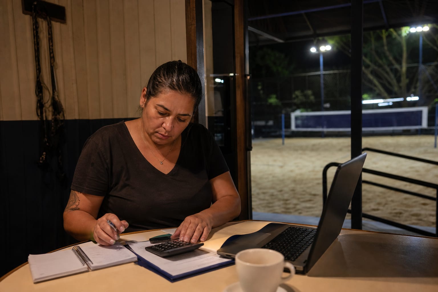 Dona de arena com calculadora, contas e notebook na recepção à noite, com a quadra iluminada visível ao fundo