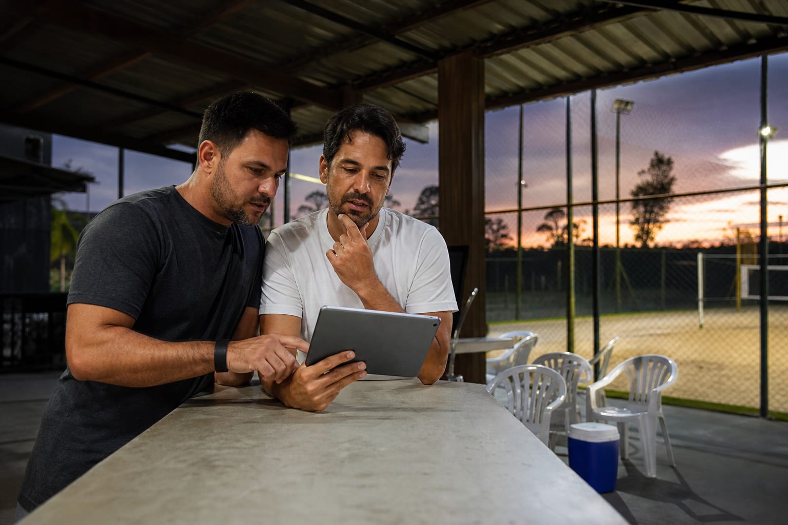 Dois sócios de arena esportiva analisando a tela de um tablet no balcão da recepção, com as quadras ao fundo